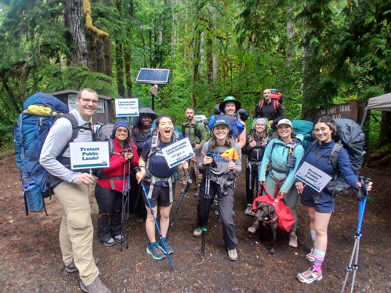 A group of smiling people with backpacks and hiking poles stand with signs that say "Protect Public Lands"