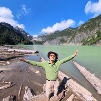 Man with glasses, plaid flannel and blue vest sits on couch with one small, white fluffy dog and a brown and tan small fluffy dog.