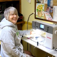 A woman sits at a sewing machine in a craft room.
