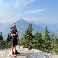 Man with blue shirt, black shirt and red backpack stands in front of lookout on a granite trail. Blue skies, evergreens and mountains in the background