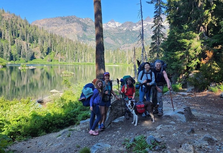 A group of three adults, two kids and a dog pose at the edge of a quiet mountain lake on a sunny morning. 