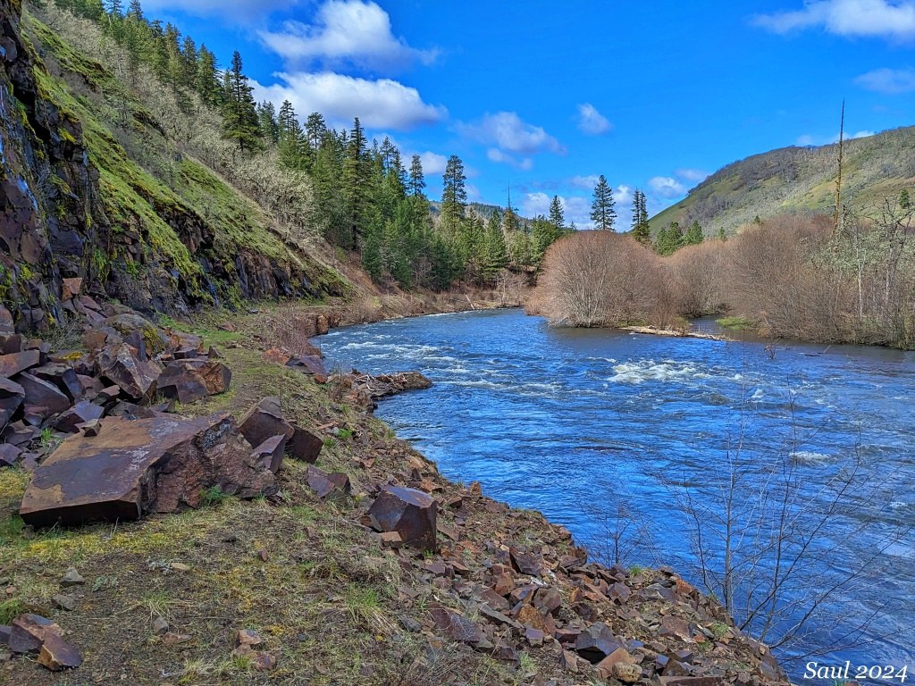 Blue river on right side of photo with foothill mountains hugging either side of water. In foreground of left hand side, red larger rocks are on the edge of water.