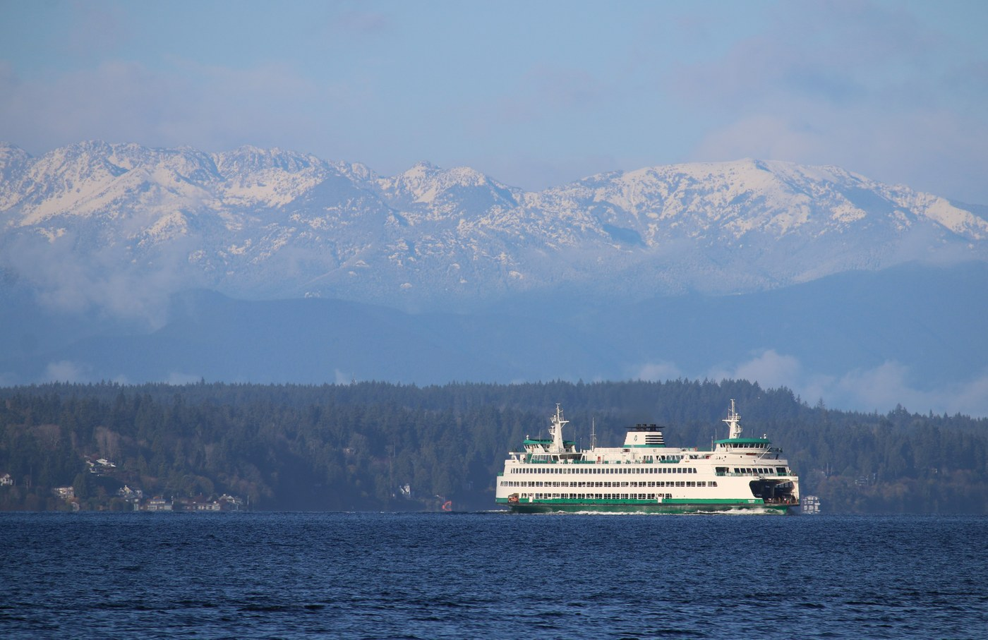 Green and White Washington State Ferry on water. Blue skies, green city foothills and snow peaked montains in the background.