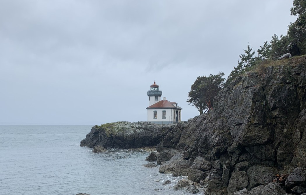 White lighthouse with red roof and blue trip stands on rocky beach. Trees line coastline on right hand side and cloudy grey sea and skies to the left.