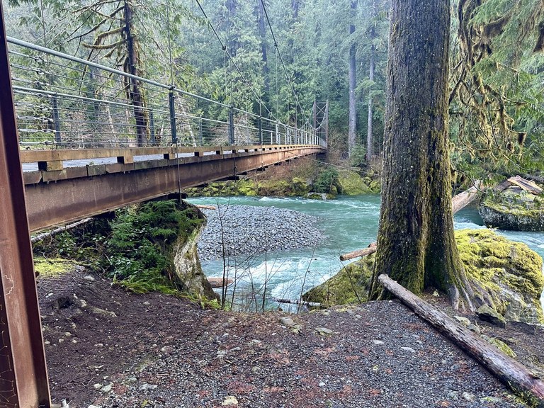A large wood bridge spans a blue-gray river lined with huge evergreens