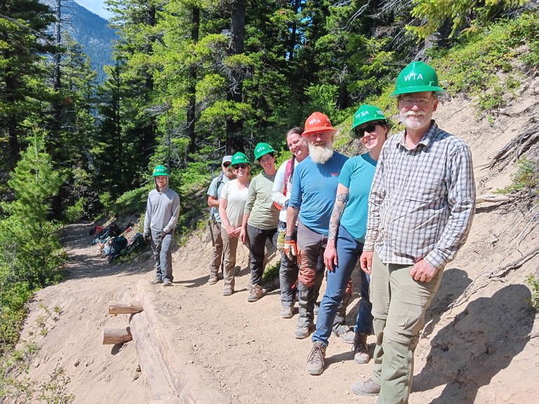 Crew smiles with hard hats on standing on a sloping dusty trail. Evergreens, mountains and blue skies in the background.
