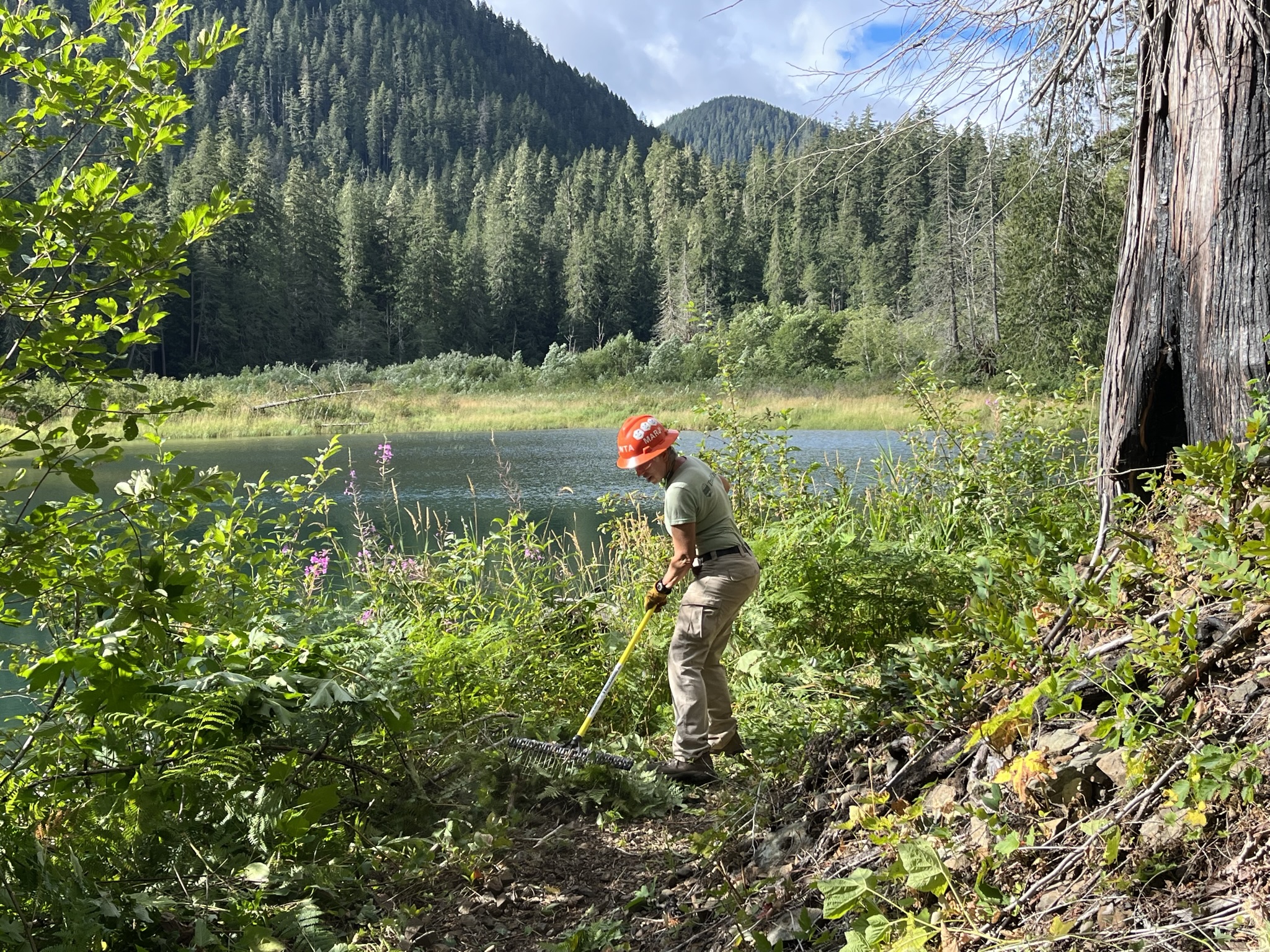 Person in hard hat works with rake beside lake and mountains.