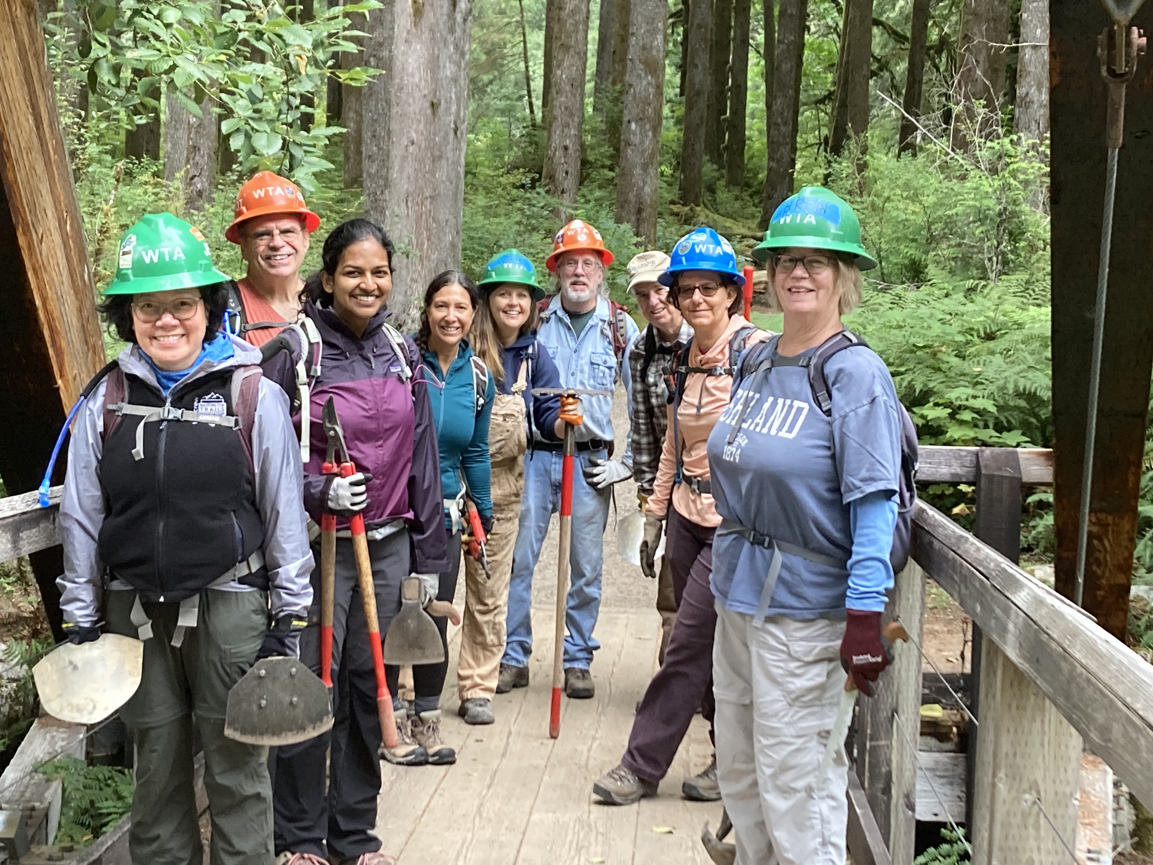 Many folks gather with trail tools in hand and hard hats on head.