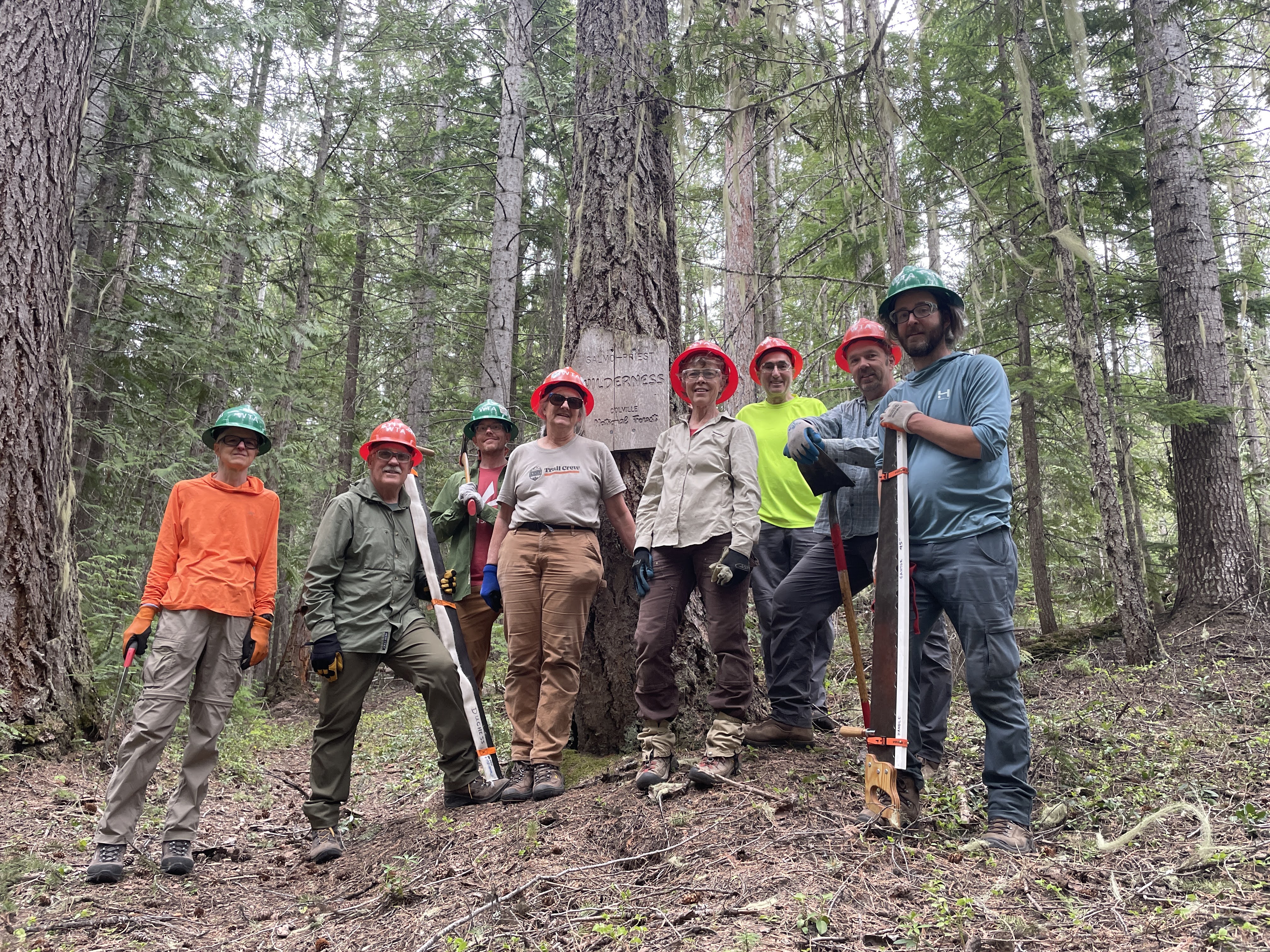 Trail crews in orange hard hats smile at camera while holding trail tools.