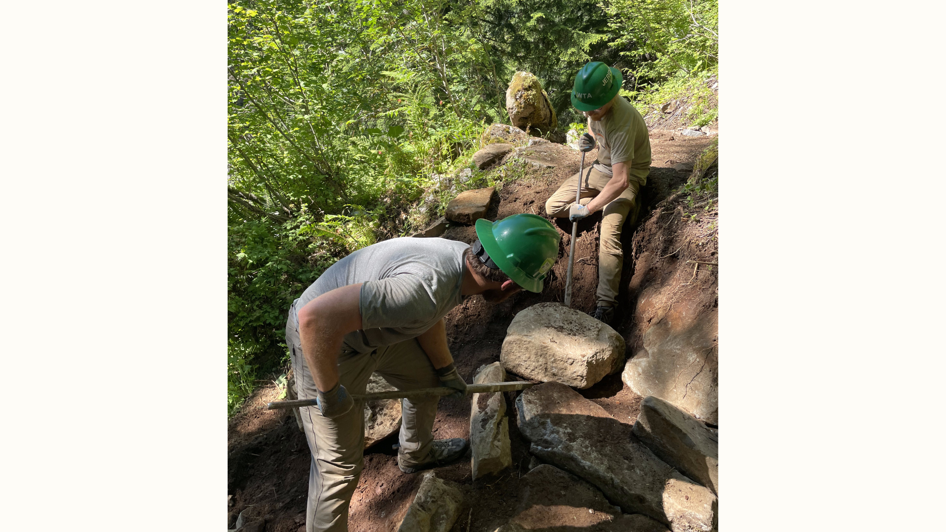 Two people in green hard hats moving a boulder to build stairs.