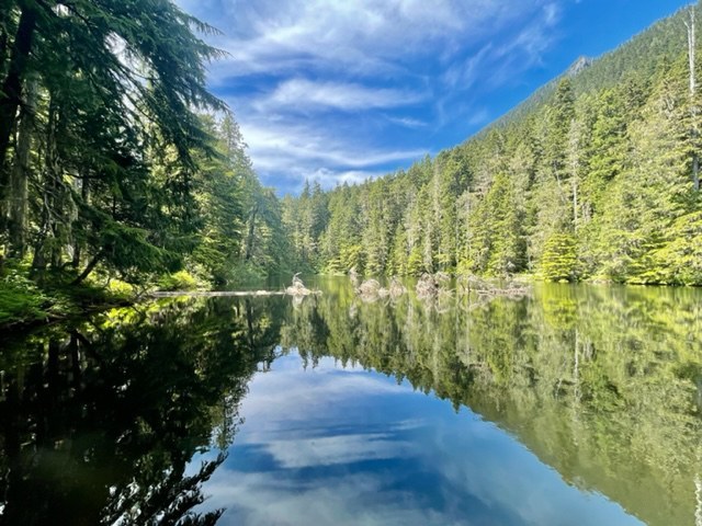 Metan Lake on a blue sky day.  A glassy lake lined with evergreen trees. The sky is bright blue with a few wispy clouds.