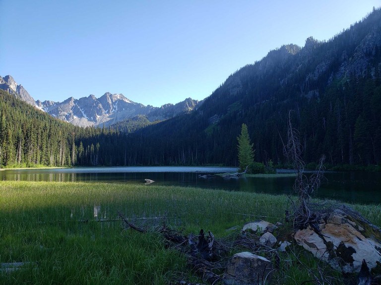A quiet alpine lake ringed by grass and high mountains is partially in shade at sunset. 
