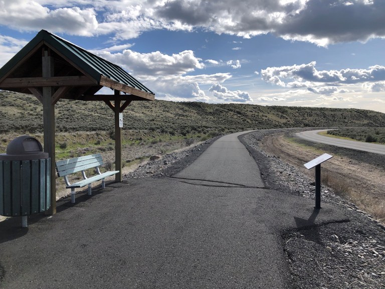 A paved trail extends to the horizon line, along with a kiosk in the foreground and a blue, cloud-scudded sky behind. 