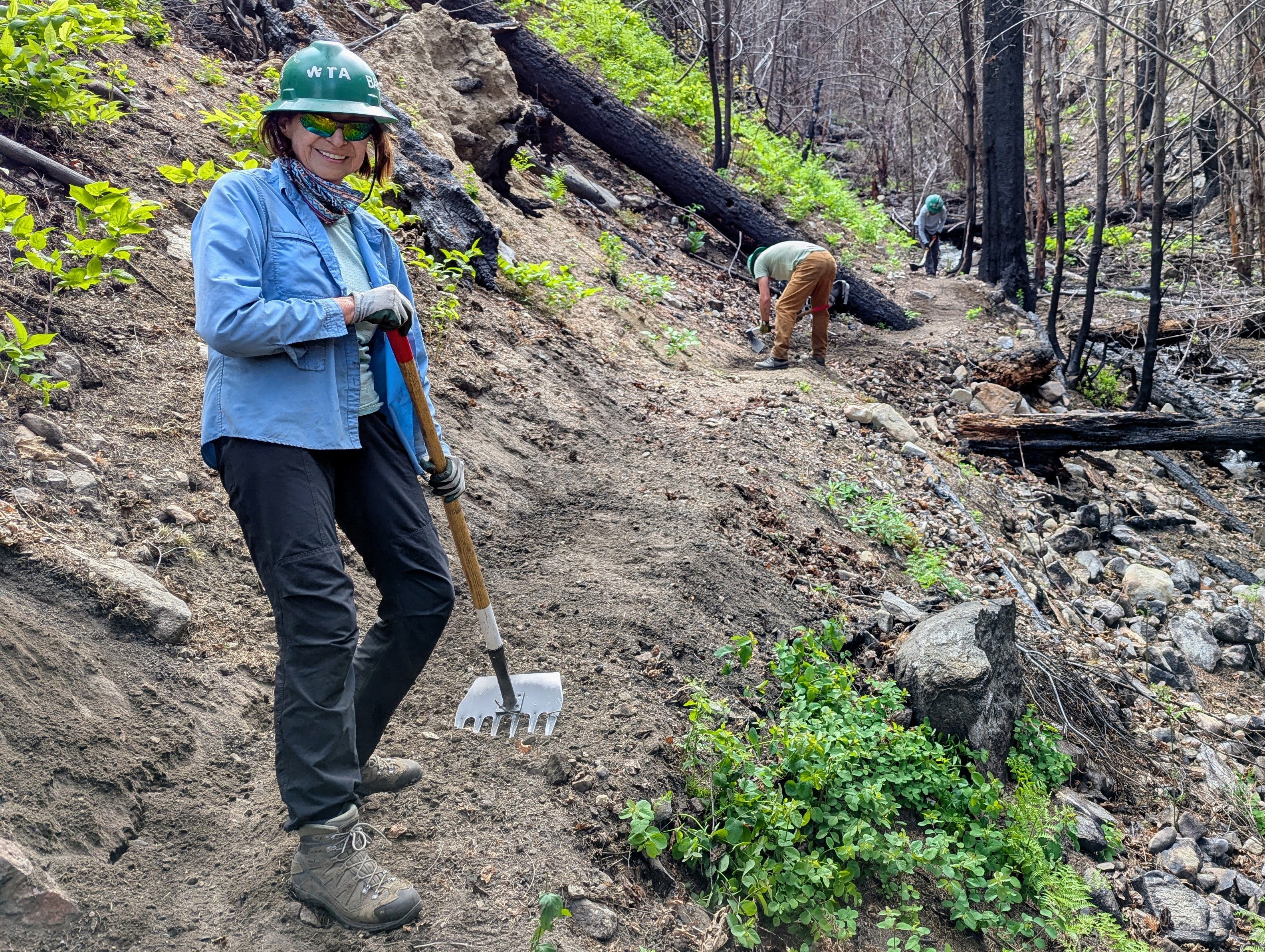 Person smiling wearing a green hard hat and holding a mcleod on a dusty trail. Burned trees and blue skies in the background. 