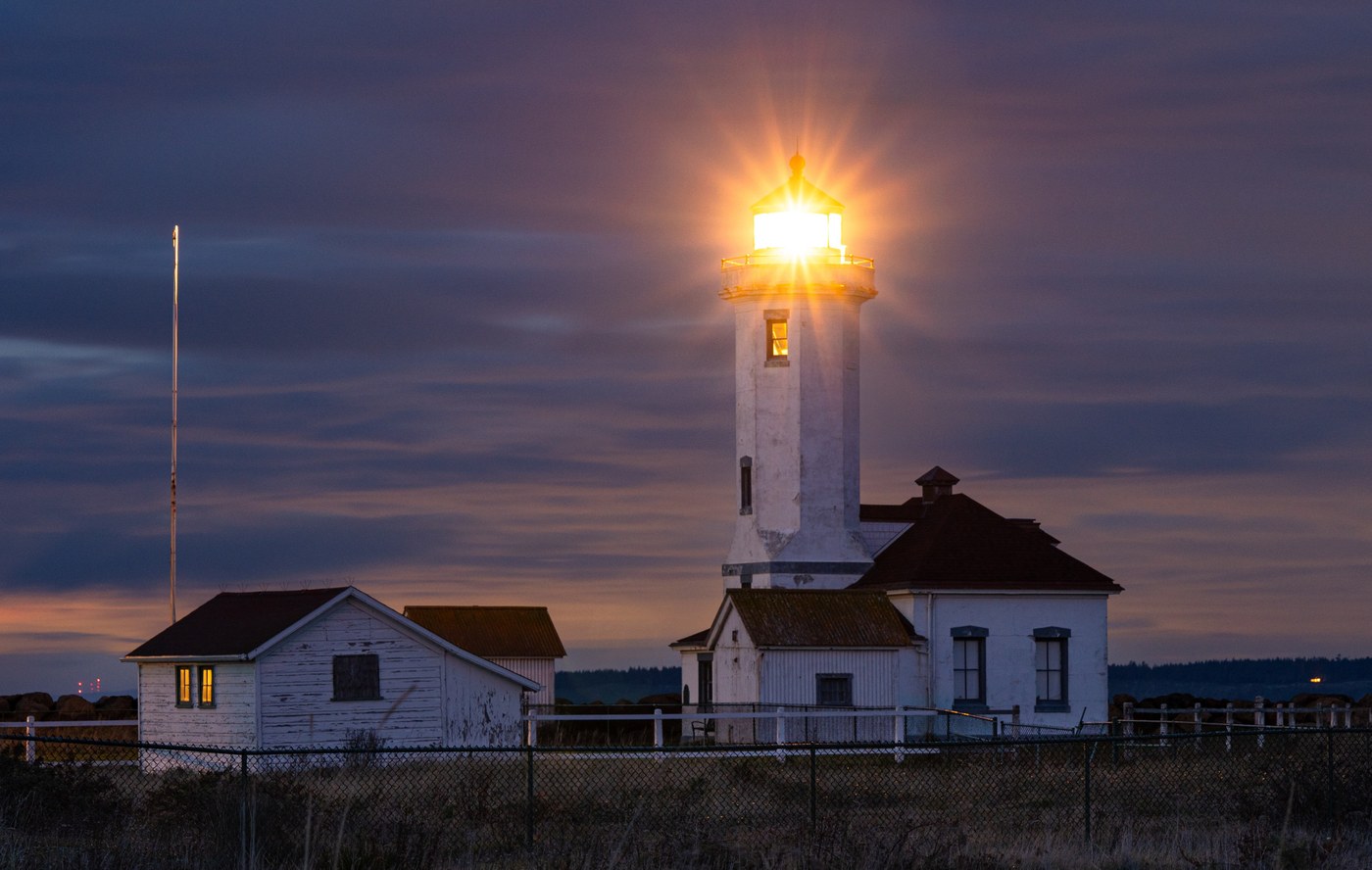The Point Wilson lighthouse emits a warm light during a purple sunset. Photo by Mancunian hiker. 