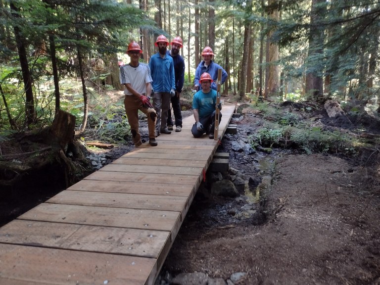 Five trail crew workers wearing orange hard hats stand at end of bridge they just built. Towering forest in the background.