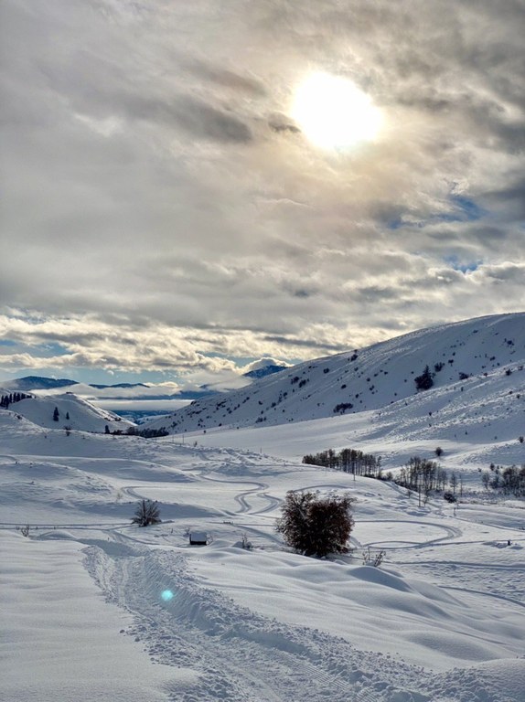 The sun glows from behind clouds as they sweep over a snow-laden landscape.