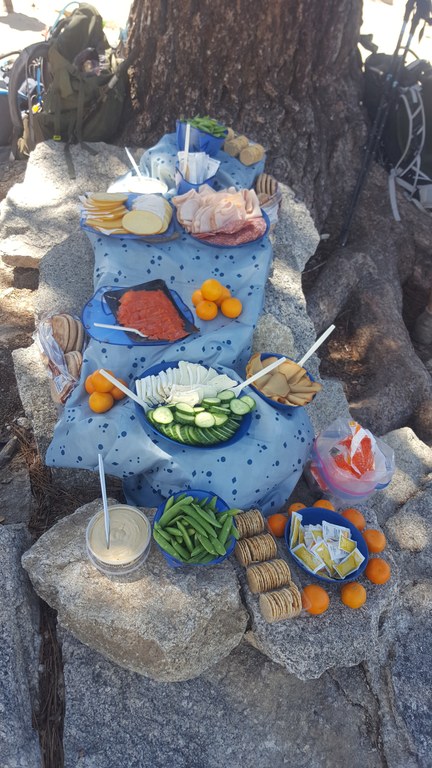 Multiple plates of food with various cheeses, meats, fruits and veggies are stacked in a cascading fashion on a tablecloth draped over rocks in the shade of a tree.