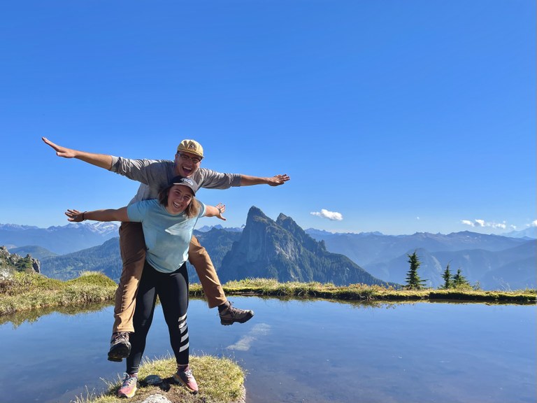 Two hikers pose with their arms out by a subalpine lake. They pose piggy-back with big smiles.