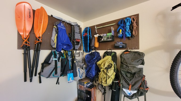 Photo by Joseph Gonzalez. A brown pegboard is hanging on a wall. It is organizing backpacks, snowshoes, kayak paddles and more.