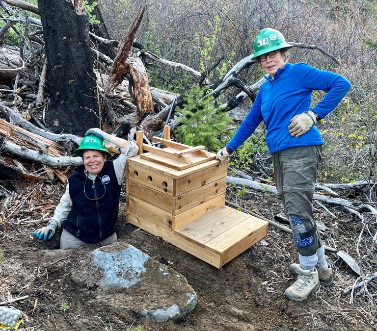 Our backcountry volunteers work hard to ensure your comfort on trail. Photo by Evelyn Hall Two WTA backcountry volunteers pose next to a newly made privy box. One of them stands inside the hole the privy will so go over.