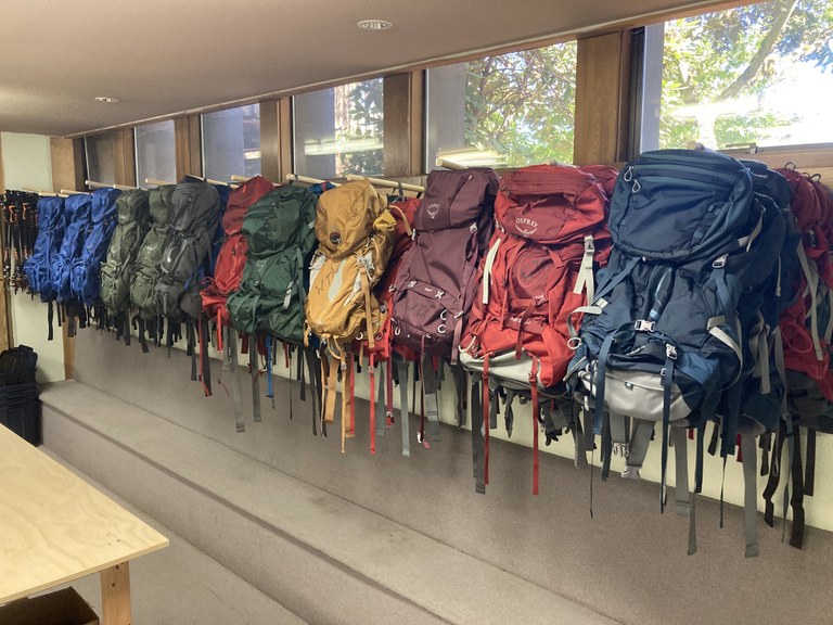 Backpacks at the WTA Puyallup Gear Library. Photo by MJ Sampang. Backpacks are hung-up against the wall in a long row. They are organized by color in a pleasing fashion.