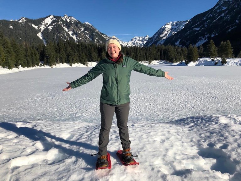 The author, wearing red snowshoes, stands in front of a frozen Gold Creek Pond with mountains and blue sky in the background. 