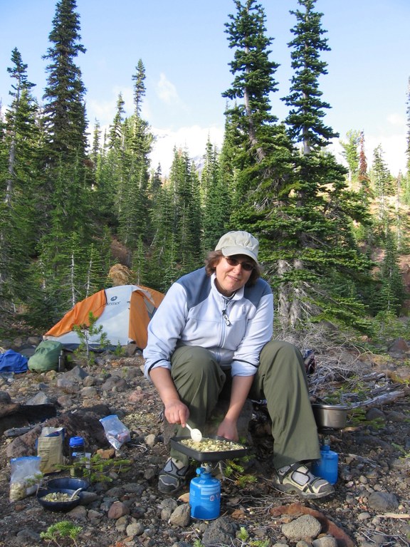 A hiker stirs dinner over a stove at camp. A tent sits in the background beneath a bushel of trees.