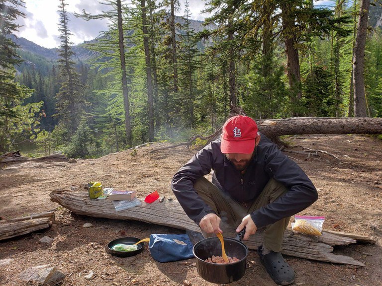 A hiker sits on a log. They're holding a pot in one hand and stirring its contents with the other.