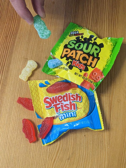 Vegan candy. Photo by Tiffany Chou. Sour Patch Kids and Swedish Fish vegan candy sitting on a table with a hand taking away a candy. Photo by Tiffany Chou.