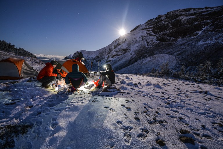 Hikers huddle around and enjoy dinner while camping on a snowy hillside.