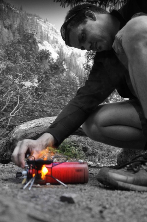 A hiker crouches over a stove as it spouts flame. The entire photo is in black and white except for the fuel canister and flame.