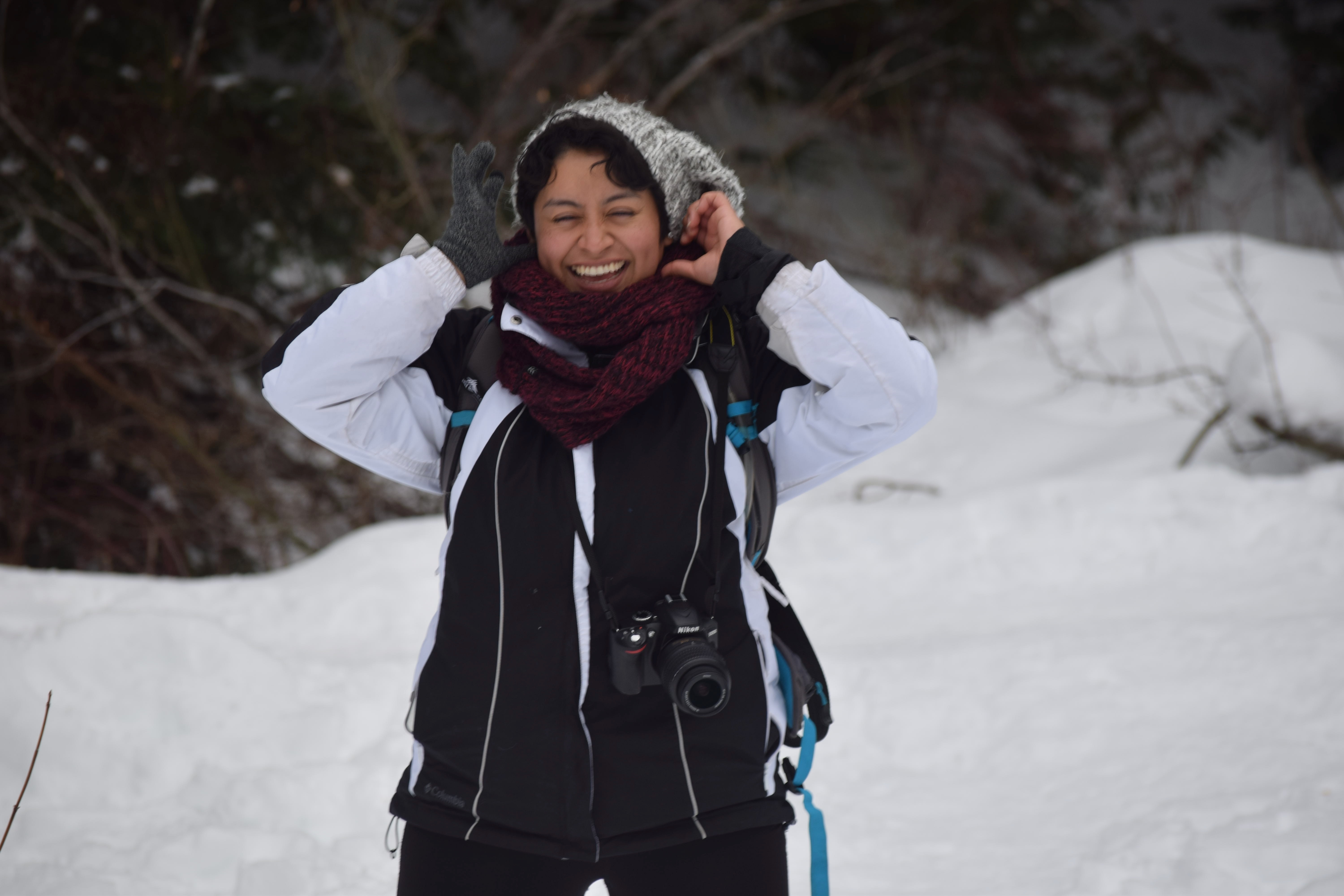 A woman stands with lots of coats on in a snowy landscape. She adjusts her hair and smiles, but is missing one glove