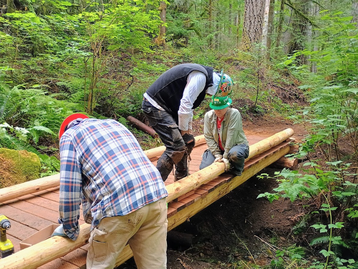 Three people in hard hats work to complete a new wooden trail bridge