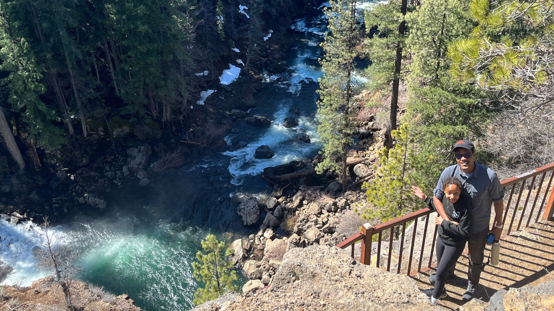 Person stands with child smiling at camera, standing in front of a railing that overlooks a rushing river below.