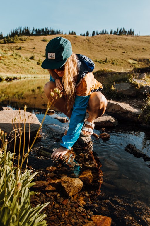 A hiker crouches on a rock in a stream. They're dipping their water bottle into the stream.