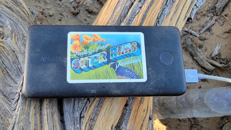 A dusty power bank sits on a piece of wood on trail. It is dusty and has a sticker on it.