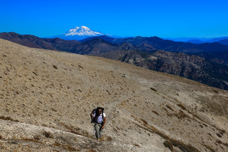 A hiker ascends a dry, steep trail. a Snowcapped volcano sits in the distance.