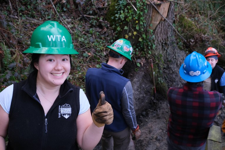 A happy volunteer working on installing new steps at Kayak Point Regional Park. Photo by Jack Hsu. One person with green helmet and thumbs up smiles at camera as two other people work in the background.