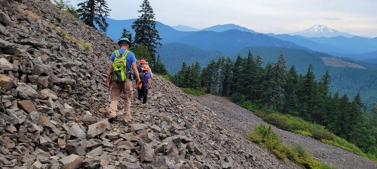 Each year, WTA’s trail maintenance program helps thousands of people make connections to each other and to special places. Photo by James Alexander Trail maintainers walk along a trail of talus. In the distance is a volcano.