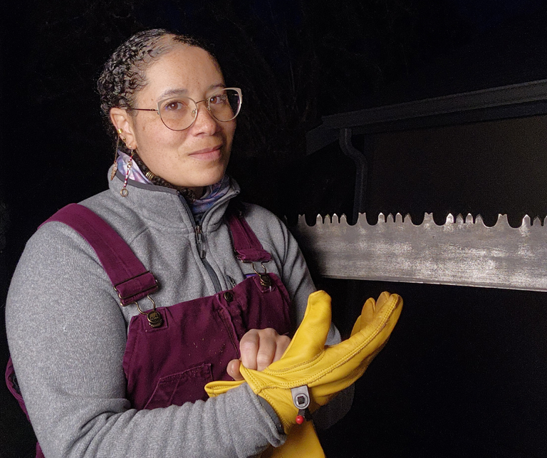 Crystal puts on work gloves while posing in front of her crosscut saw.