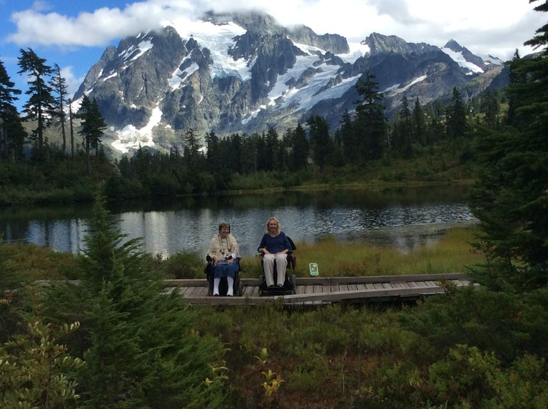 Two hikers using wheelchair smile at the camera in front of a large picturesque lake with a snow capped mountain in the background.