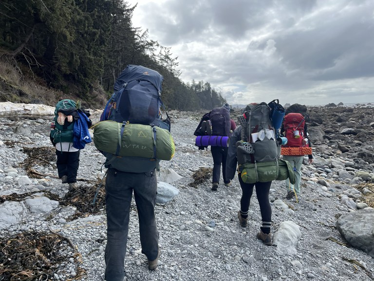 Five backpackers hike along a pebbly stretch of trail, right next to some tidepools.