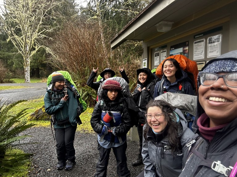 Hikers smile and pose at the trailhead at Olympic National Park. They're wearing rain gear and backpacks, but smile ear to ear.