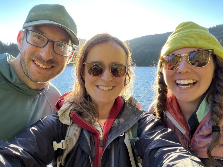 Three hikers smile at a lake.
