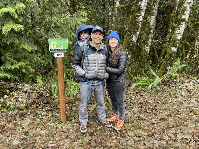 Jaime Loucky and his family. Jaime Loucky and his family stand at the trailhead of the Olympic Discovery Trail. They are wearing jackets, hats, and hiking shoes. Jaime's child sits in a backpack on his back.