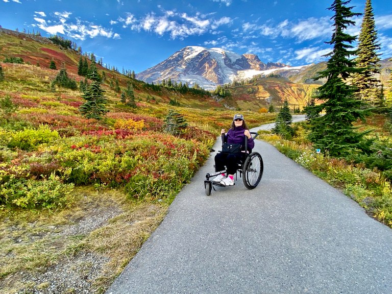 Jenny on a paved section of trail on Mount Rainier, fall colors in the meadows on either side and the mountain in the distance.