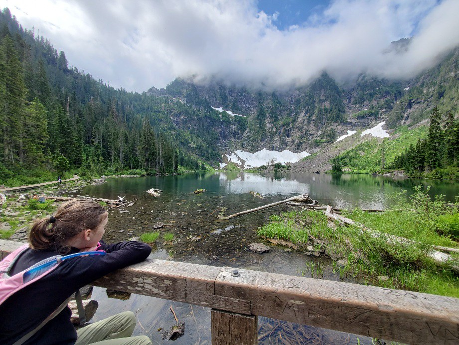 A young hiker with a ponytail and pink backpack leans on a railing and gazes out over the lake basin