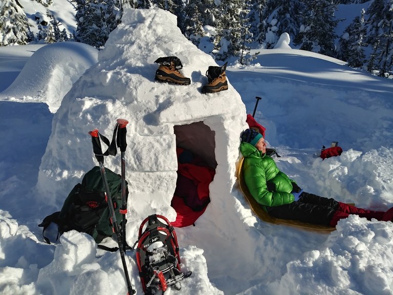 A hiker sits in the snow and enjoys some sun. Their back is to a snow structure, which they've built. On top of the snow structure are various articles of clothing.