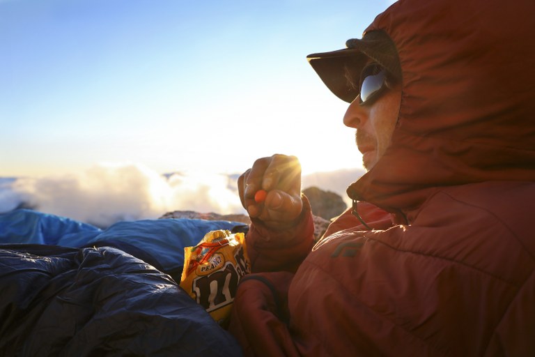 A person sits in their sleeping bag while wearing a jacket. They are enjoying chocolate candy while looking at a beautiful view.